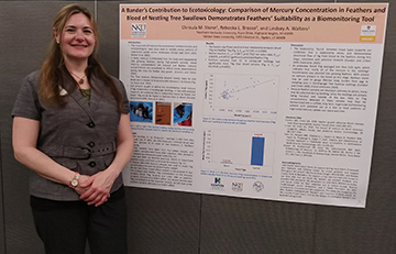 Chrisula Stone stands beside her wall-mounted poster smiling